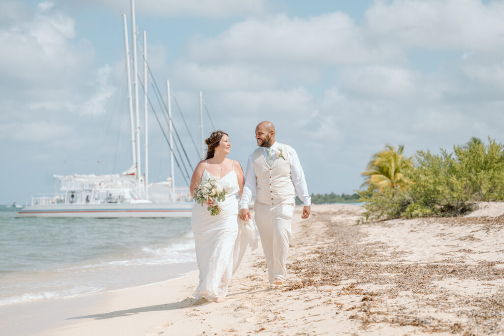 A newly married couple walk along a beach in Cozumel with sailboats in the back