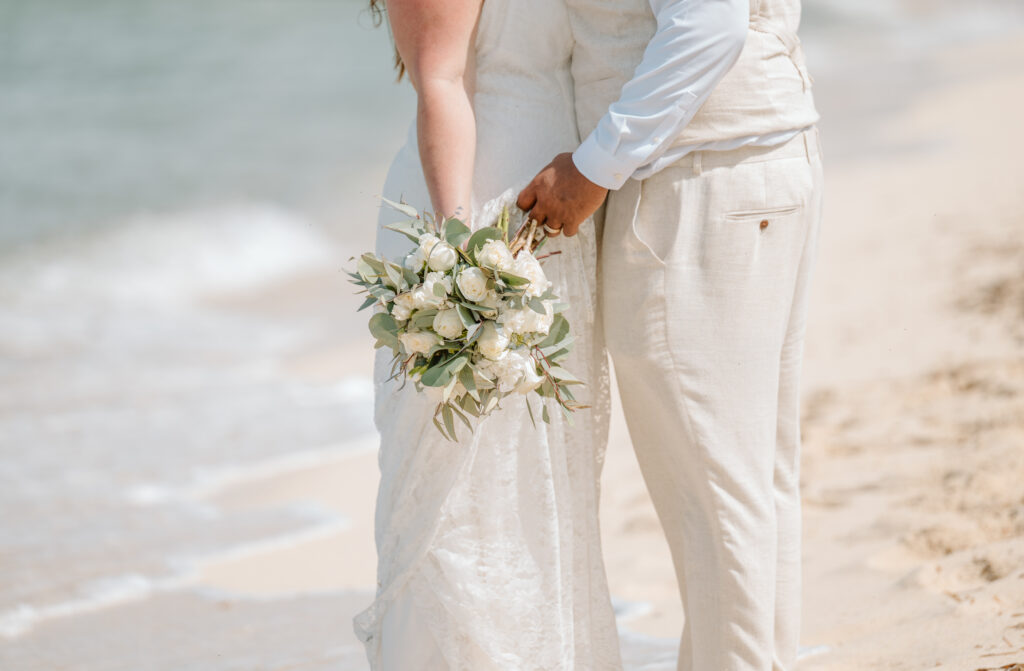 A groom and bride hold a white rose bouquet near the beach