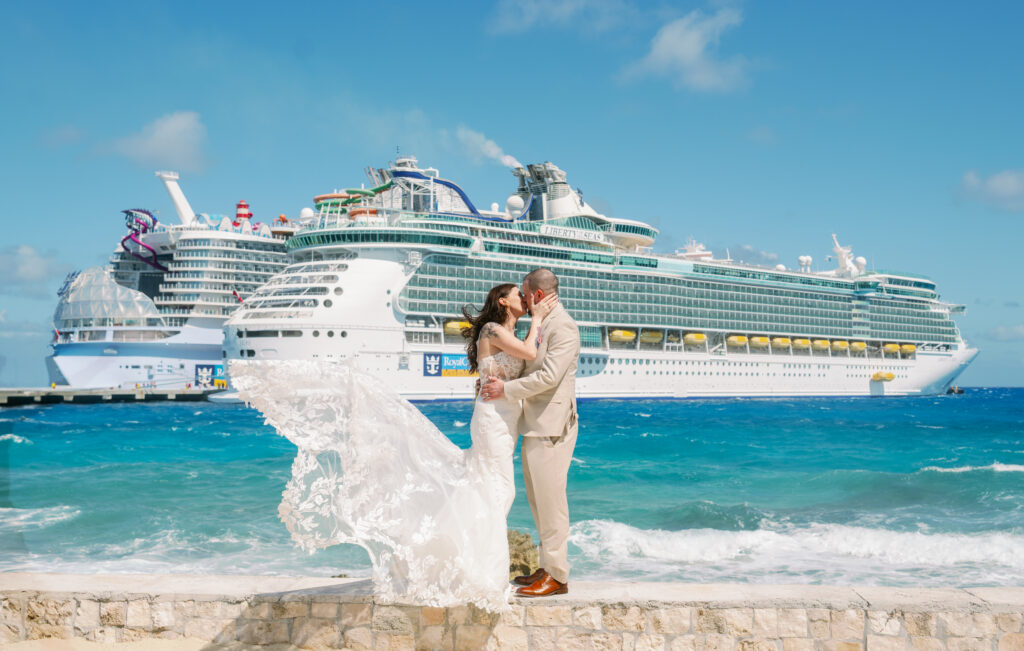 A groom in a tan suit kisses his bride while her dress blows in the wind in front of two cruise ships in the Caribbean